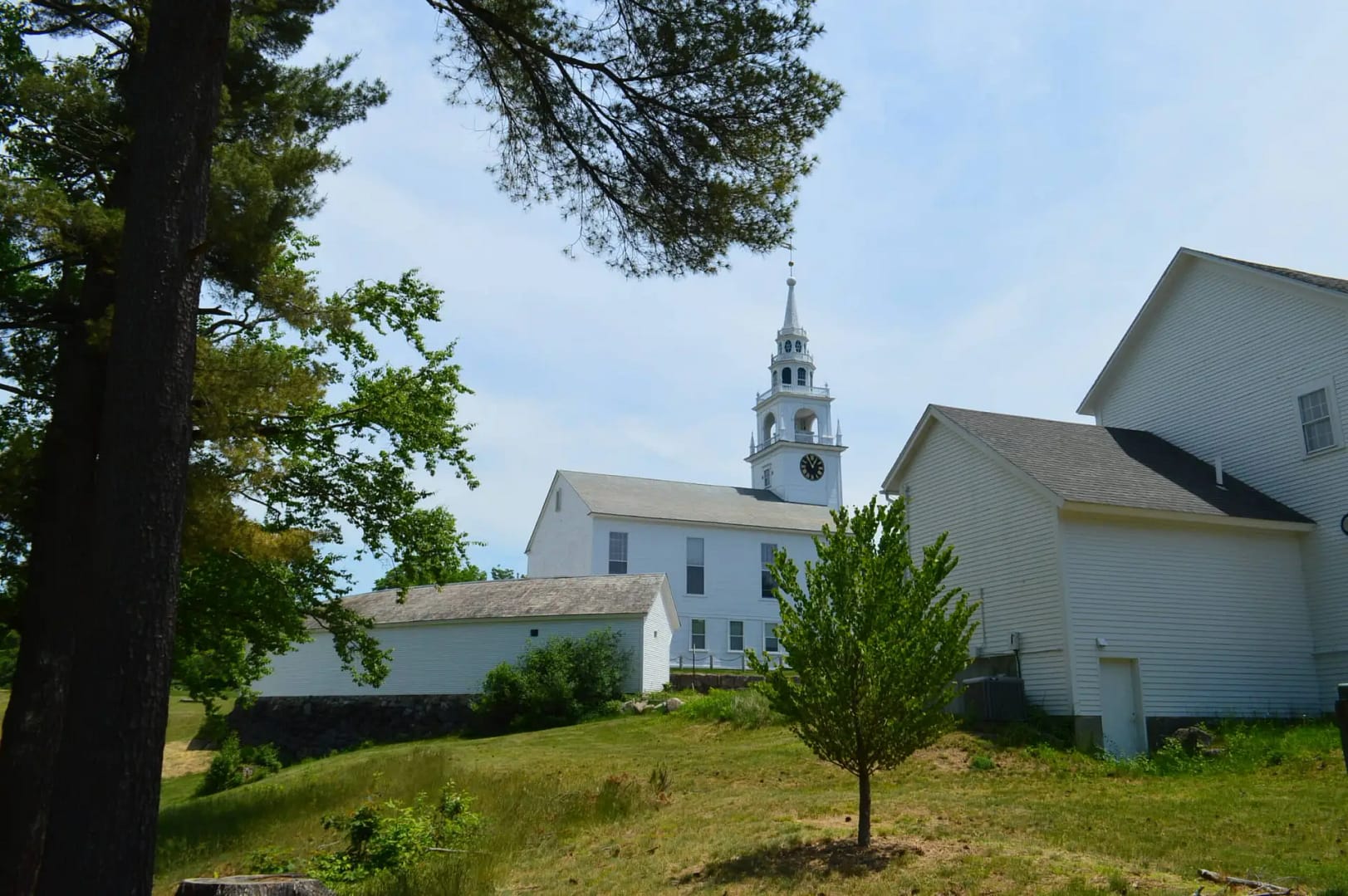 A white church with a clock tower stands on a grassy hill in Hancock NH, surrounded by trees and other white buildings under Spacious Skies.