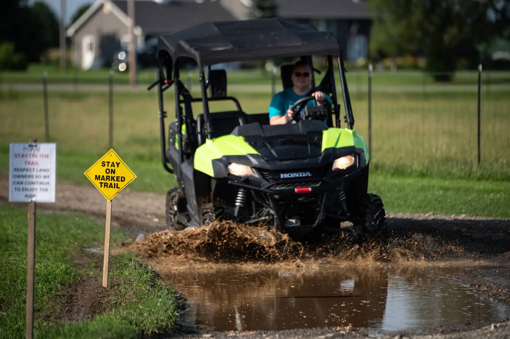 A person driving a green Honda utility vehicle through a muddy puddle on an off-road trail. A yellow diamond-shaped sign beside the trail reads "Stay on marked trail," and a white sign with smaller text is partially visible in the foreground. Houses are in the background.