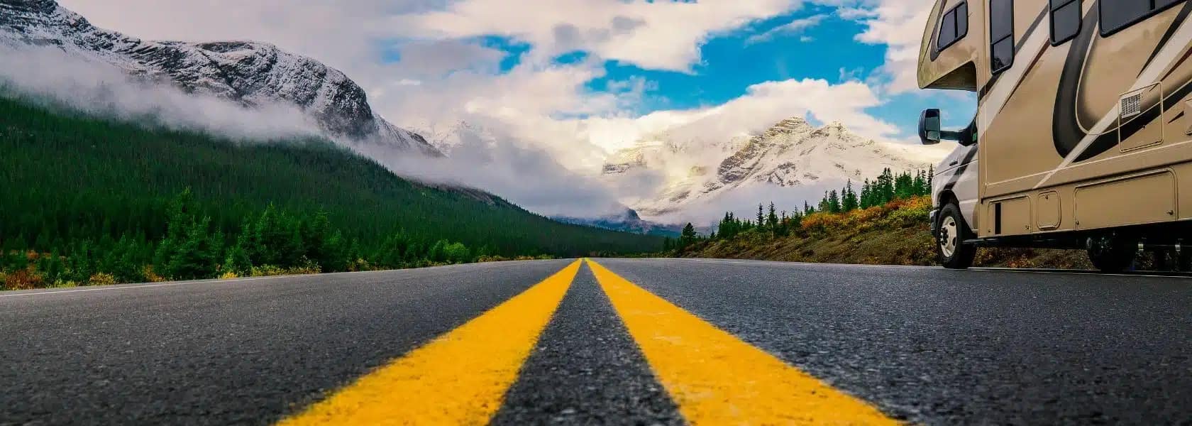 A motorhome is parked on a road leading towards snow-capped mountains and forests under a partly cloudy sky at the Spacious Skies Campground.