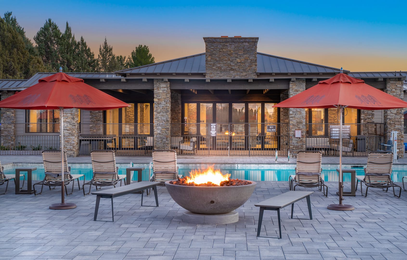 Outdoor pool area with lounge chairs, red umbrellas, and a lit fire pit in front of a stone clubhouse at sunset.