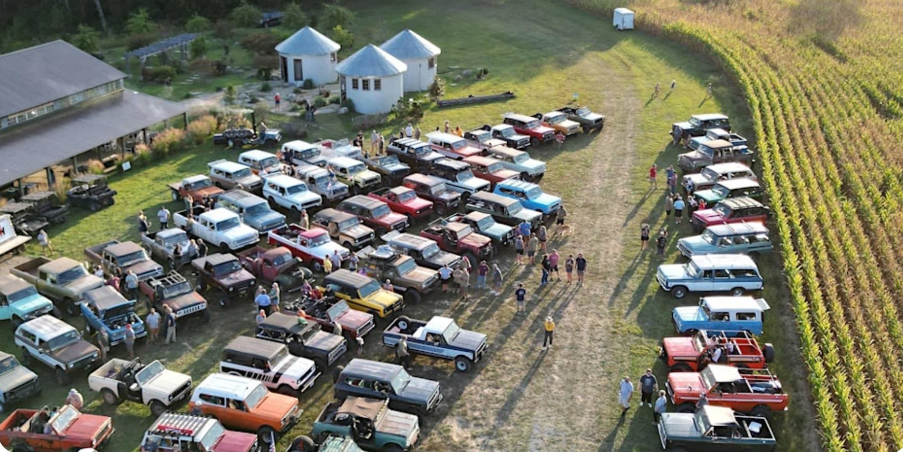 Aerial view of a grassy field with many classic trucks, SUVs, and harvesters parked in rows. People are walking and gathering around the vehicles, with buildings, a cornfield, and the bustle of the holler in the background.