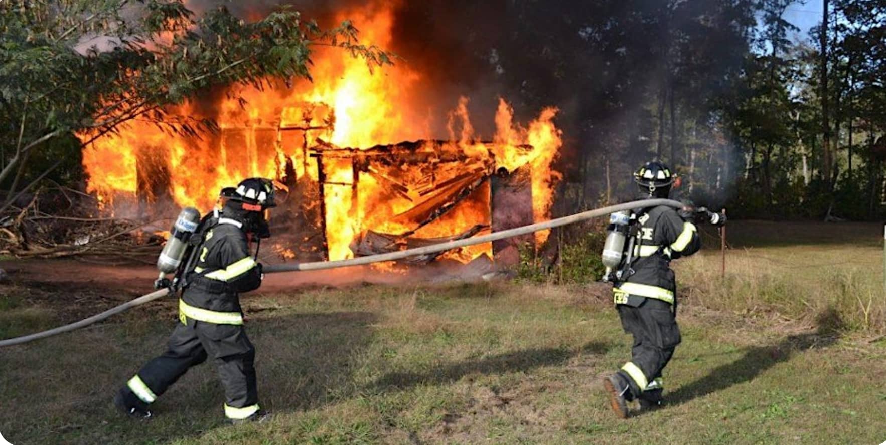 Two firefighters in full gear from Upper Cumberland Fire carry a hose toward a house engulfed in large flames, surrounded by grass and trees under a clear sky.