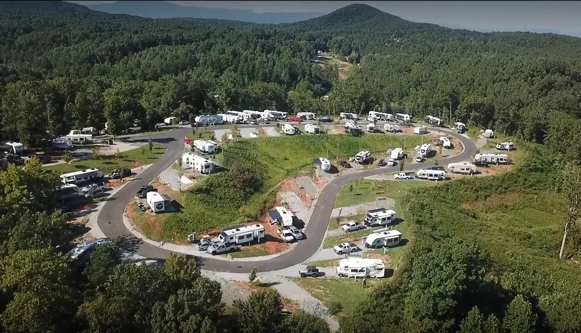 An aerial view of a campground reveals numerous RVs and trailers parked on curved roads, nestled in a dense green forest with distant mountains framing the scene. Find your comfort zone amidst nature's splendor at this tranquil retreat.