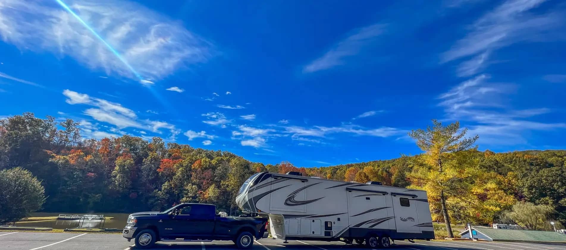 A pickup truck towing a large fifth-wheel rv travels through an area with colorful autumn foliage under a clear blue sky.