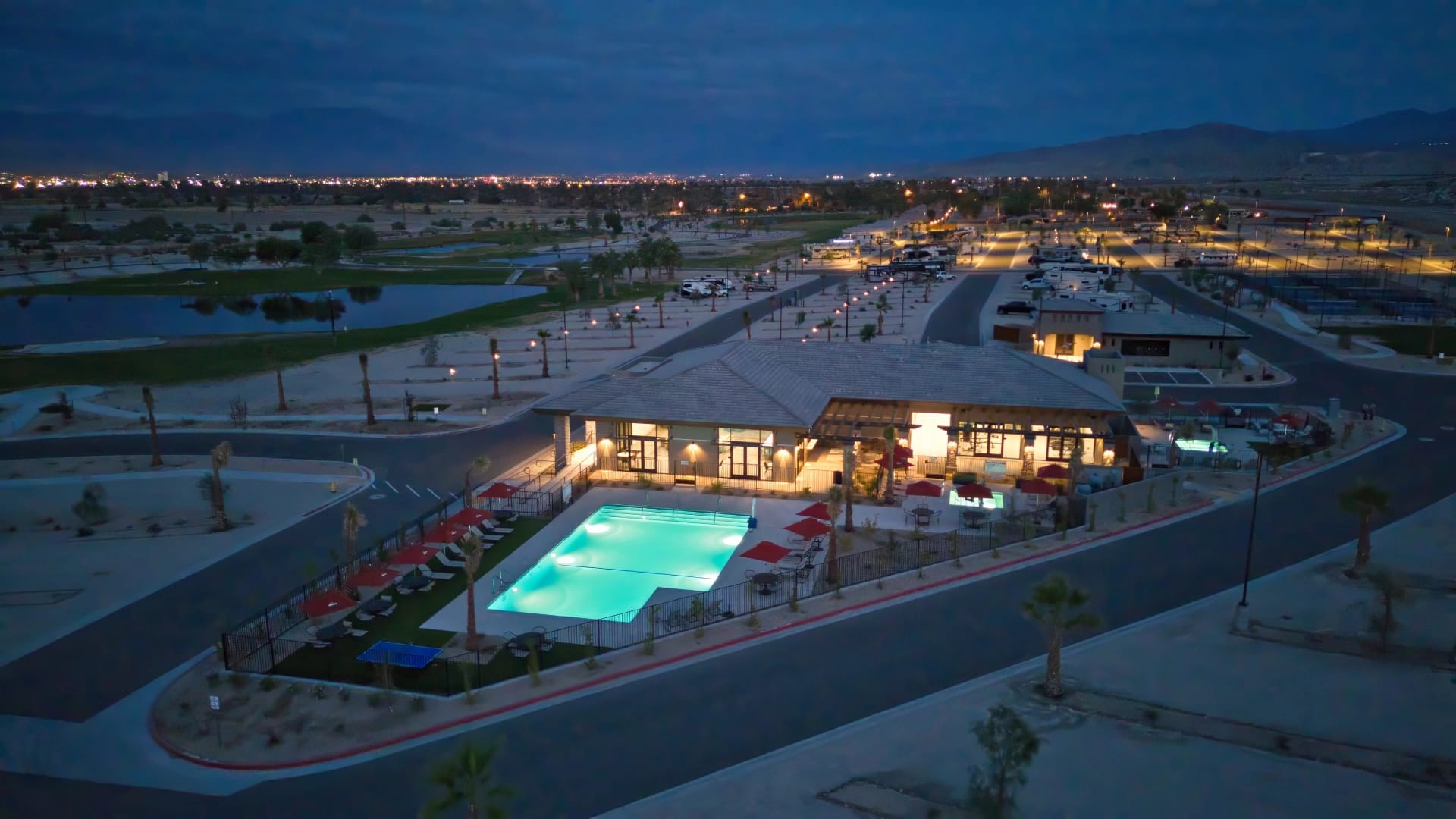 Aerial view of a brightly lit swimming pool and clubhouse at night, surrounded by parking lots, roads, and a desert landscape with city lights in the distance.