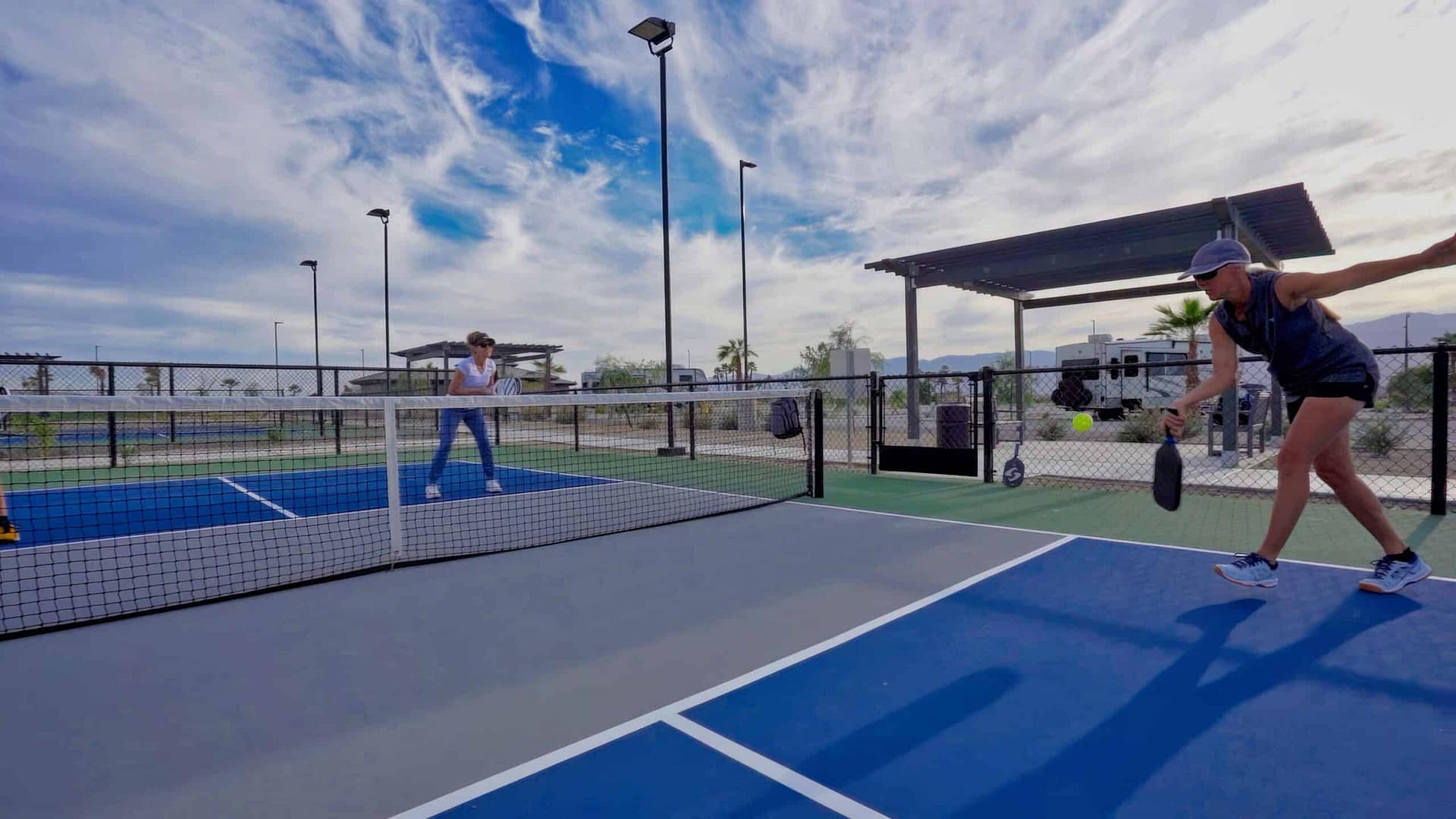 Two people play pickleball on an outdoor court under a partly cloudy sky. One player prepares to hit the ball while the other stands near the net.