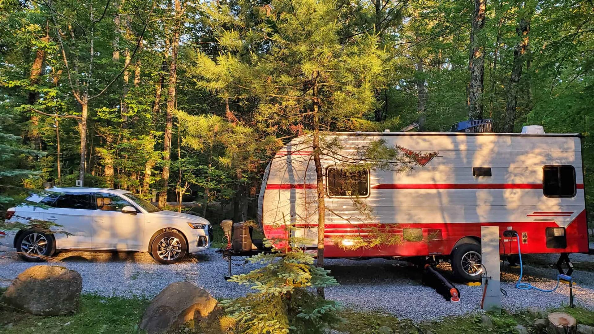 An RV parked next to a car in the spring woods, surrounded by blooming flowers along a campground trail.