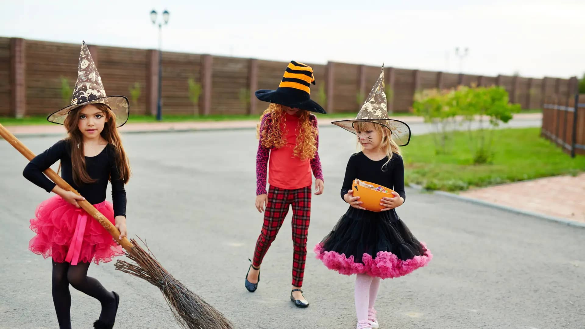 Three children wearing witch hats and colorful outfits stroll down a street in Woodland Hills. One holds a broom, another sports a large striped hat, and the third carries an orange bucket, perhaps for the Haunted Halloween Fest scavenger hunt. A fence and some greenery are visible in the background.