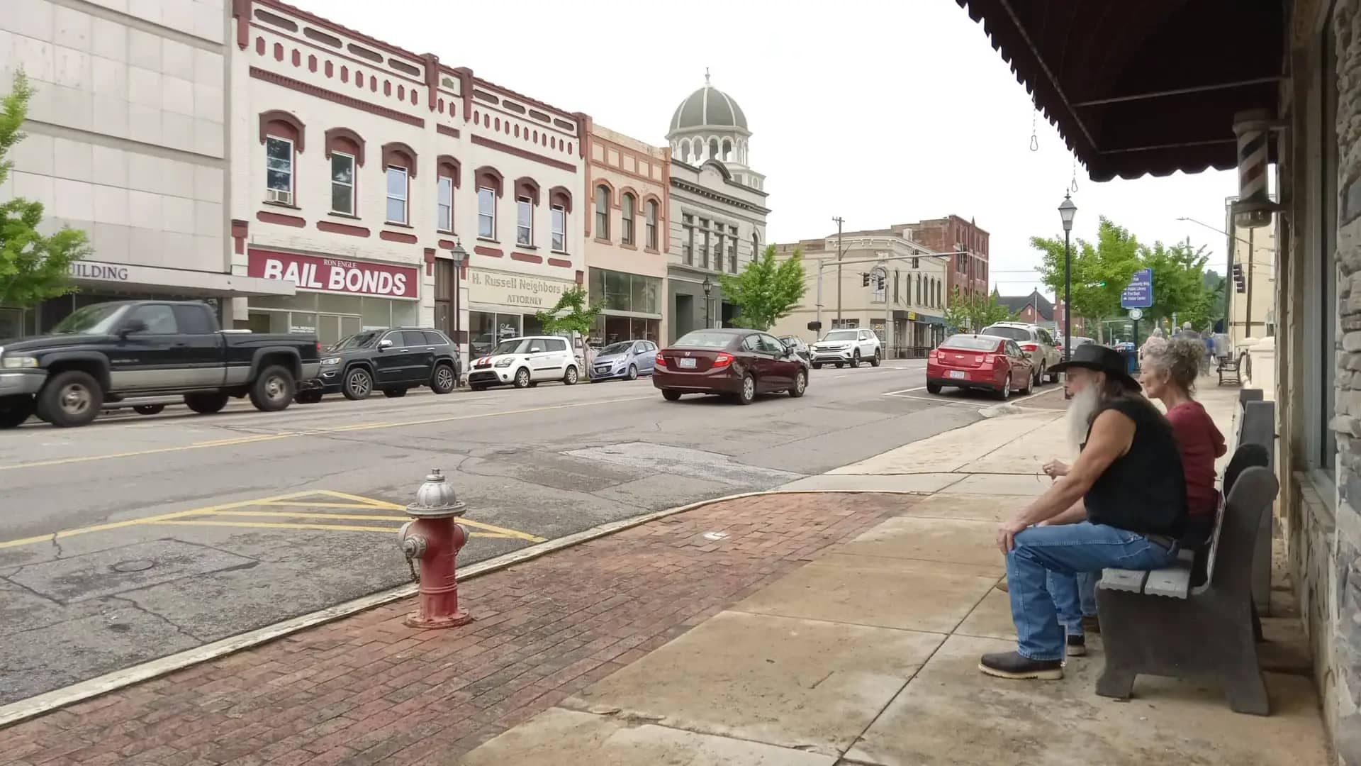 Two people sit on a bench on a sidewalk in a small downtown area with parked cars, old buildings, and a courthouse dome visible in the background. They sip wine and smile, surrounded by planters brimming with wildflowers that bring an added touch of charm to the picturesque scene.