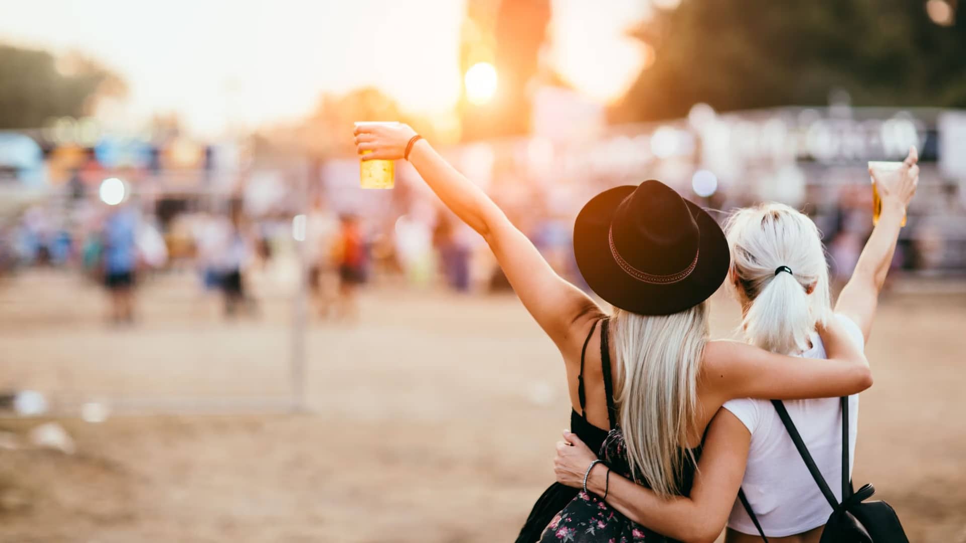 At the Tybee Post Music Festival, two people embrace, raising their drinks in celebration. One sports a black hat, while the other carries a backpack. As the sun sets, it casts a warm glow over the vibrant festival crowd welcoming Music Festival 2024 with open arms.