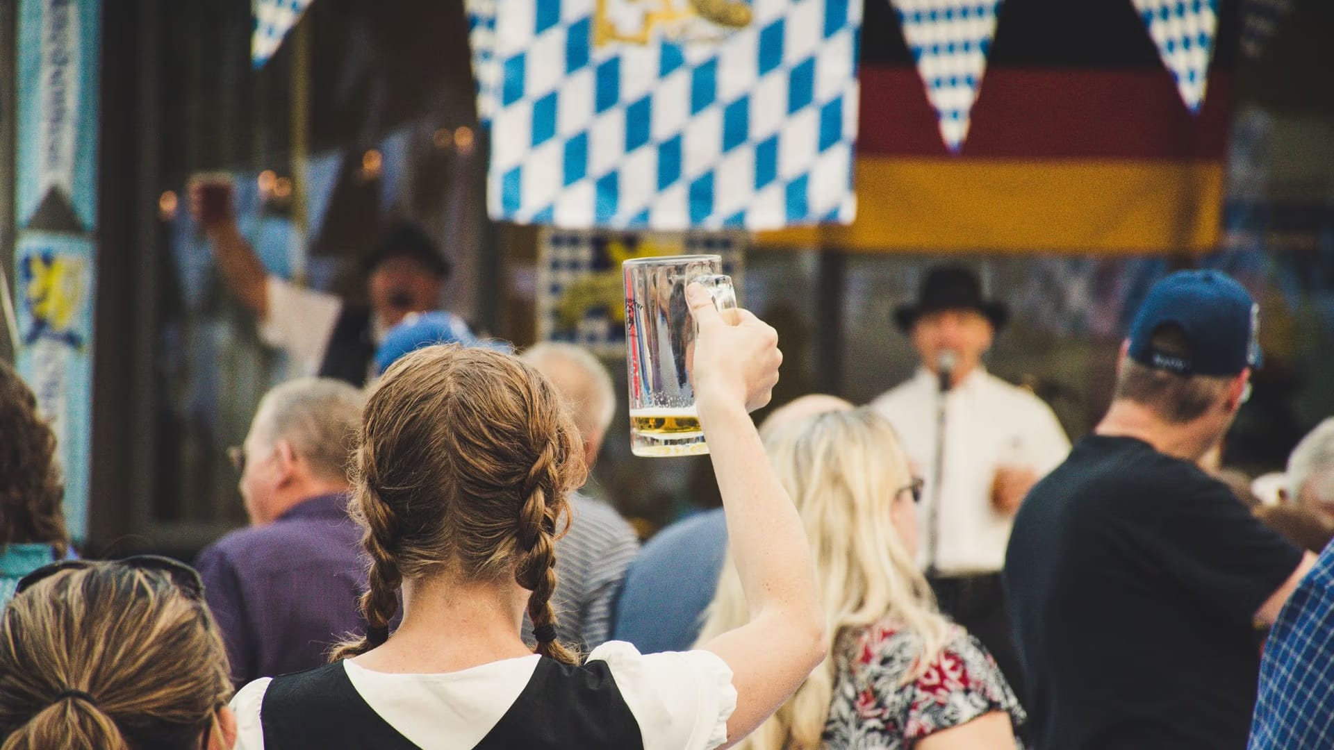 Amidst a vibrant Oktoberfest celebration at Plant Riverside District, a woman in the foreground raises a large glass of beer. Blue and white checkered banners are strung above, while two performers in traditional outfits captivate the crowd from the stage in the background.