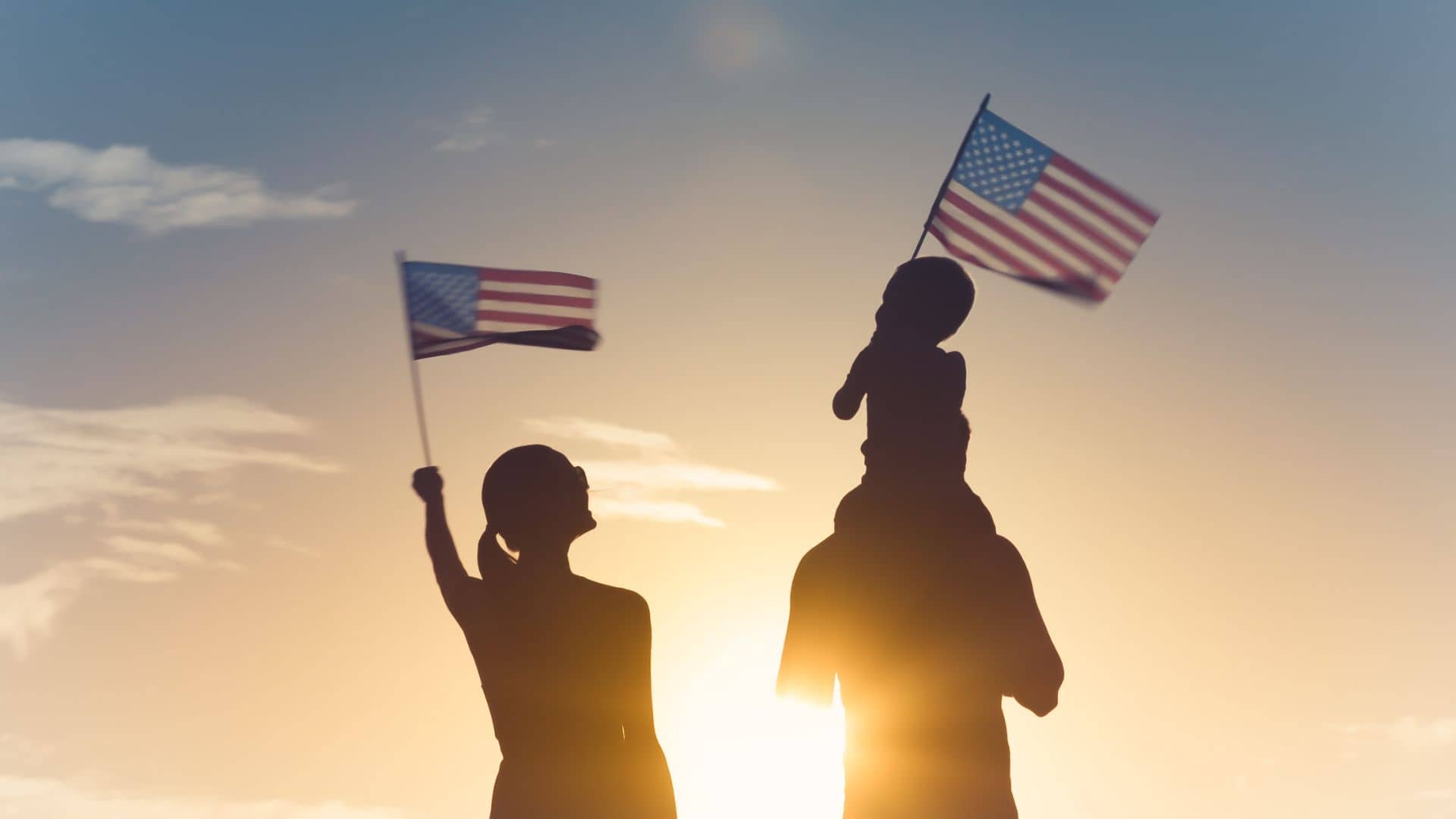 Silhouettes of a family against a sunset, with a child sitting on an adult's shoulders, embody the spirit of military families. Both adults and the child wave American flags in the clear sky at Spacious Skies Sandy Run Campground, creating an ideal getaway for cherished moments together.
