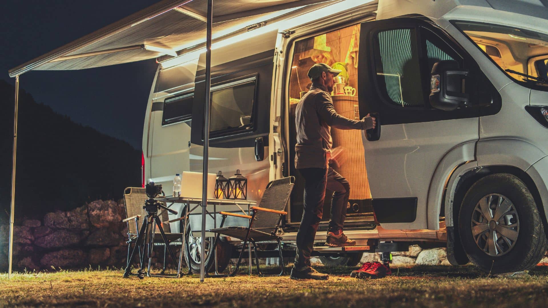 A person stands by an illuminated camper van at night, holding a mug under the starlit sky of Belle Ridge Campground in TN. The van's open door and extended awning reveal cozy camping chairs and a small table, perfect for winter camping nights with a softly glowing lantern nearby.
