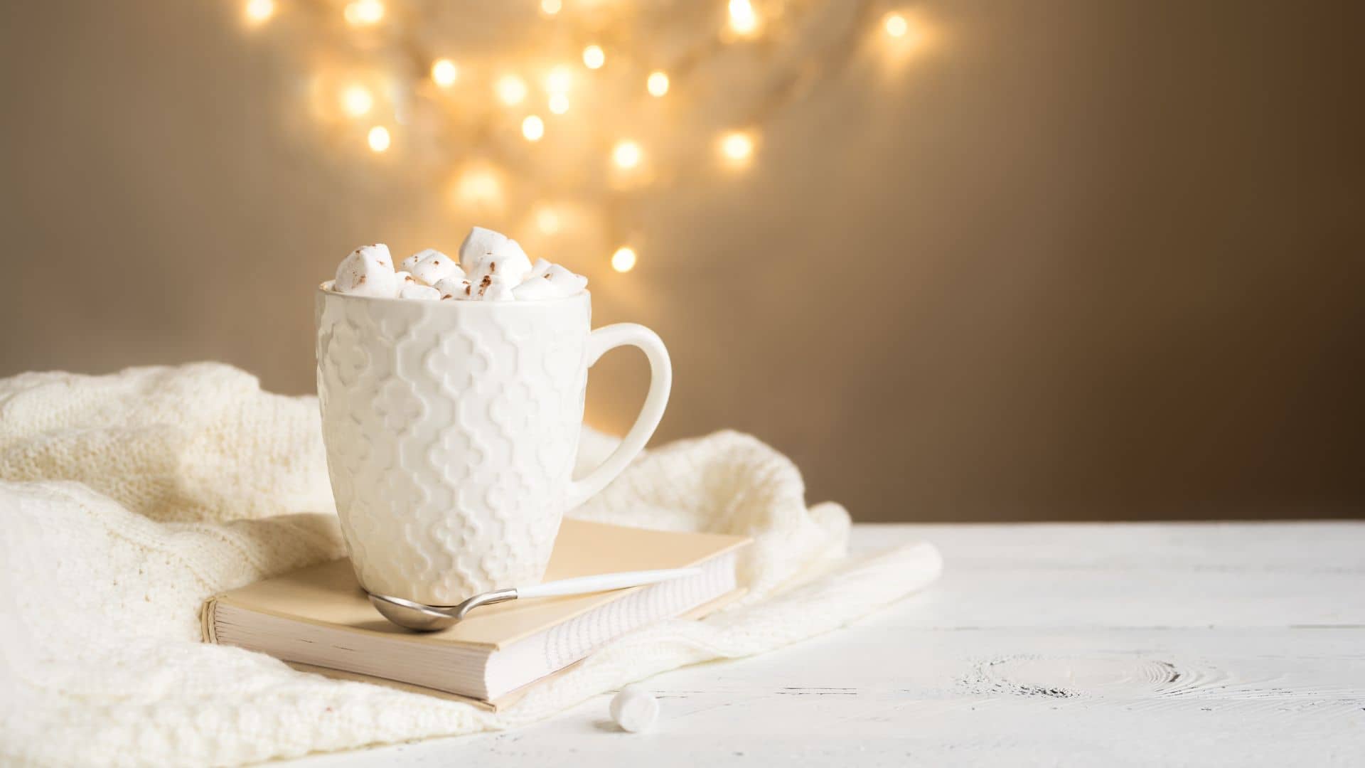 A white mug filled with hot chocolate and marshmallows sits on a closed book with a spoon nearby. Soft, glowing lights and a cozy knitted blanket complete the scene, creating the perfect camping space that embodies a hygge-inspired warmth and inviting charm.