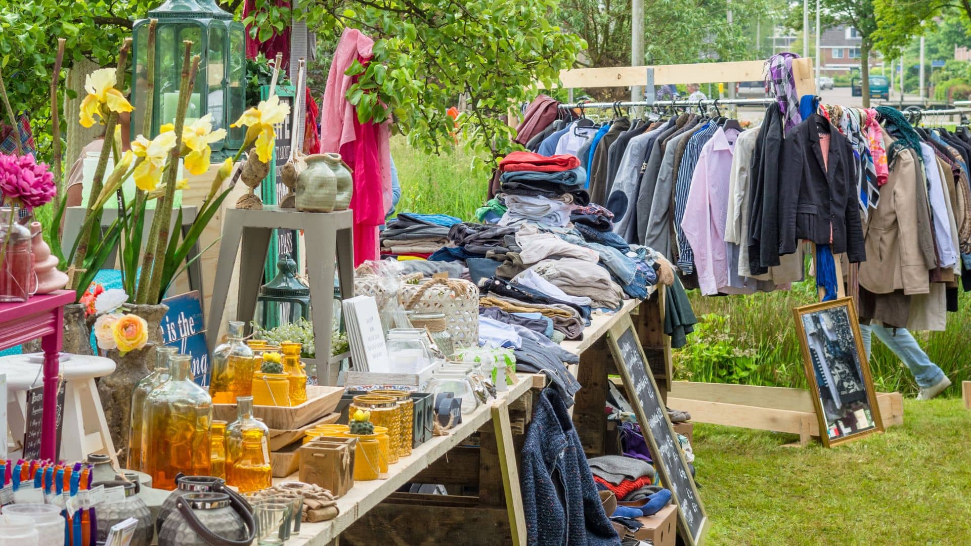 At a bustling spring sidewalk yard sale, a vibrant outdoor flea market stall showcases various items. Clothes are piled on tables and hung on racks, while glass bottles, vases, and decorative objects glint in the sun. Lush greenery envelops the area, crafting an inviting atmosphere.