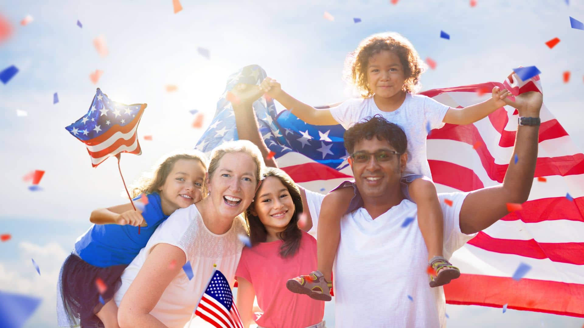 A diverse family of five celebrates outdoors in Downtown Marion with smiles, holding American flags. Two children wave flags as one child sits on an adult's shoulders, surrounded by festive red, white, and blue confetti under the clear 4th of July sky.