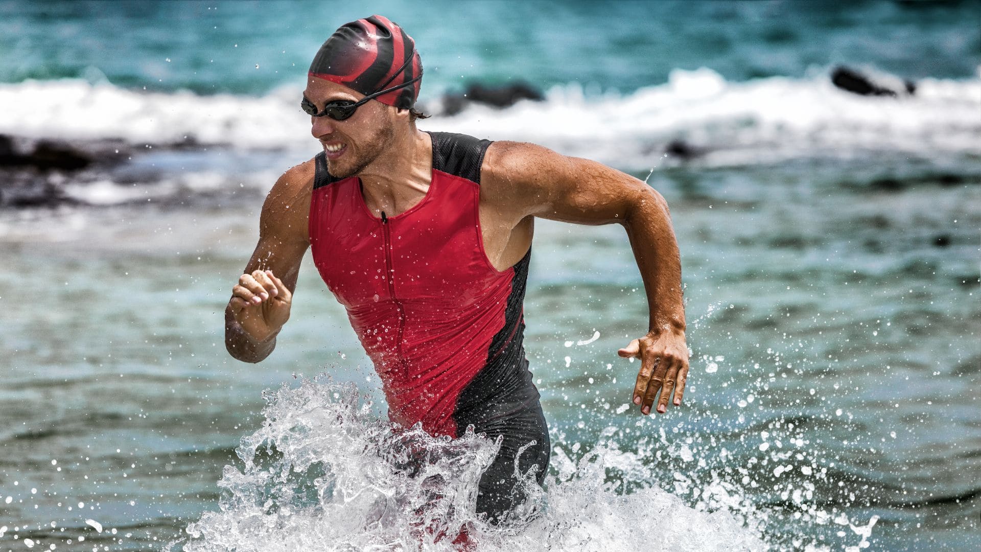 A person wearing a red and black triathlon suit, swim cap, and goggles is emerging from the ocean, running through splashing water. This Ironman competitor mirrors the determination seen in Lake Placid events. The background shows waves and a blue sea under a clear sky.