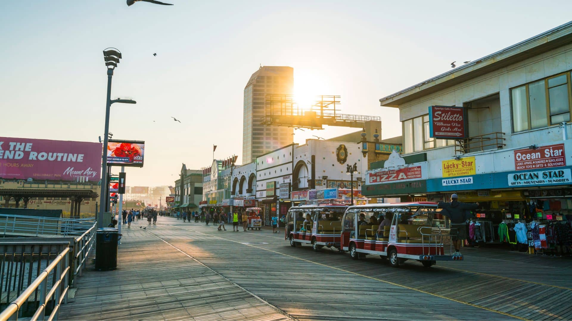 A lively boardwalk scene features shops and eateries with bright signs, perfect for a springtime adventure. People stroll along as a red sightseeing tram passes by. The sun sets behind a tall building, casting a warm glow over the area, enhancing the charm of this vibrant destination.
