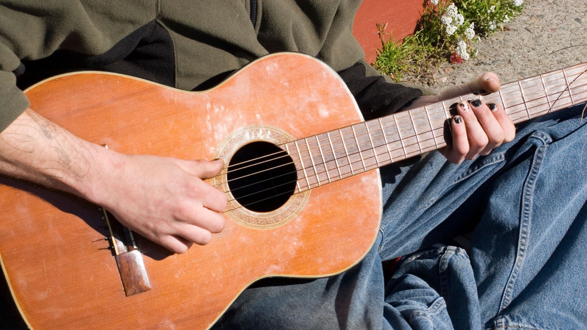 A person strums an acoustic guitar outdoors during 4th Friday in Downtown Fayetteville, wearing a dark jacket and blue jeans. Their fingernails are painted black, and small white flowers are scattered on the ground nearby.