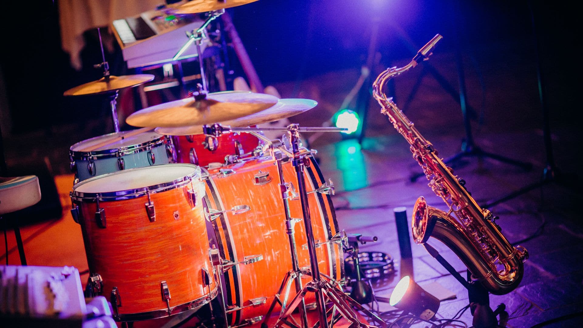 A drum set and a saxophone grace the dimly lit stage, ready to ignite a festival vibe. The drum set boasts a glossy orange finish, while the saxophone stands poised beside it. Colorful stage lights cast a vibrant glow, setting the perfect scene for unforgettable music moments.