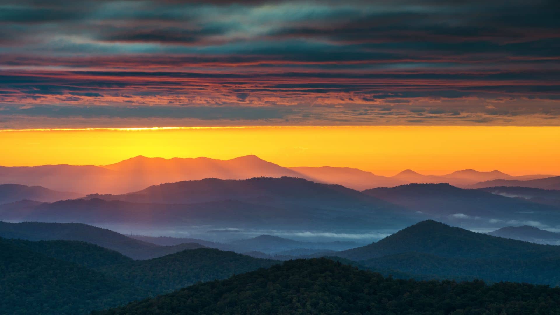 A scenic mountain landscape at sunrise with layers of misty blue hills, a vibrant orange-yellow horizon, and dark, dramatic clouds above. Sunbeams peek through, casting light and shadows over the terrain.