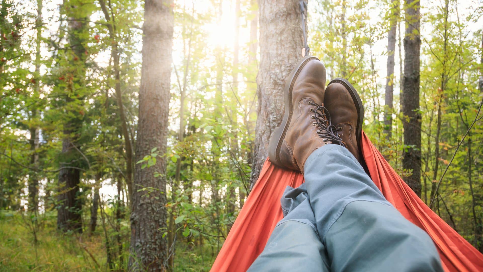 Person relaxing in a hammock in a forest, wearing brown boots and blue-gray pants. Sunlight filters through the trees, creating a peaceful and calm atmosphere.