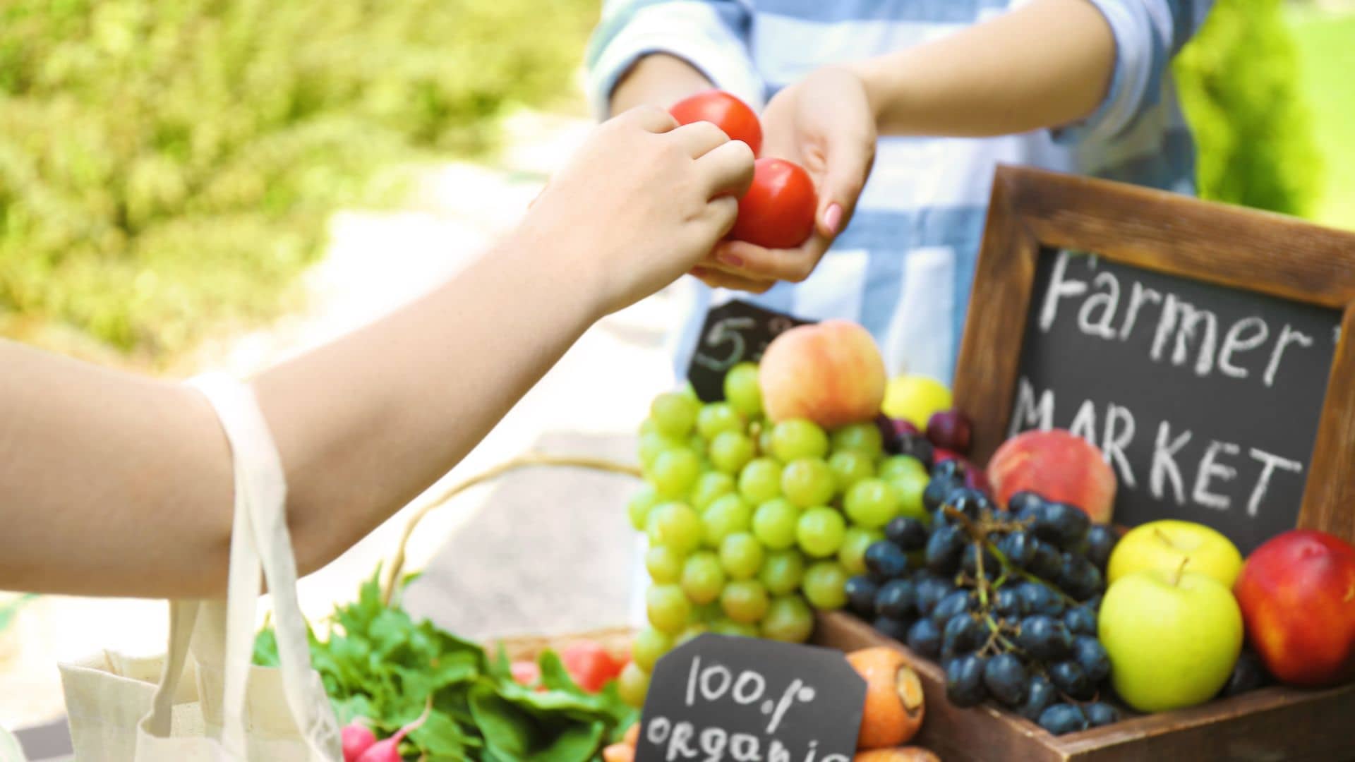 A person buys tomatoes from a stall at Hancock's Farmers Market. The stall boasts baskets of fresh produce like grapes, apples, and greens. Charming chalkboard signs proclaim "100% organic" at this delightful artisans market.