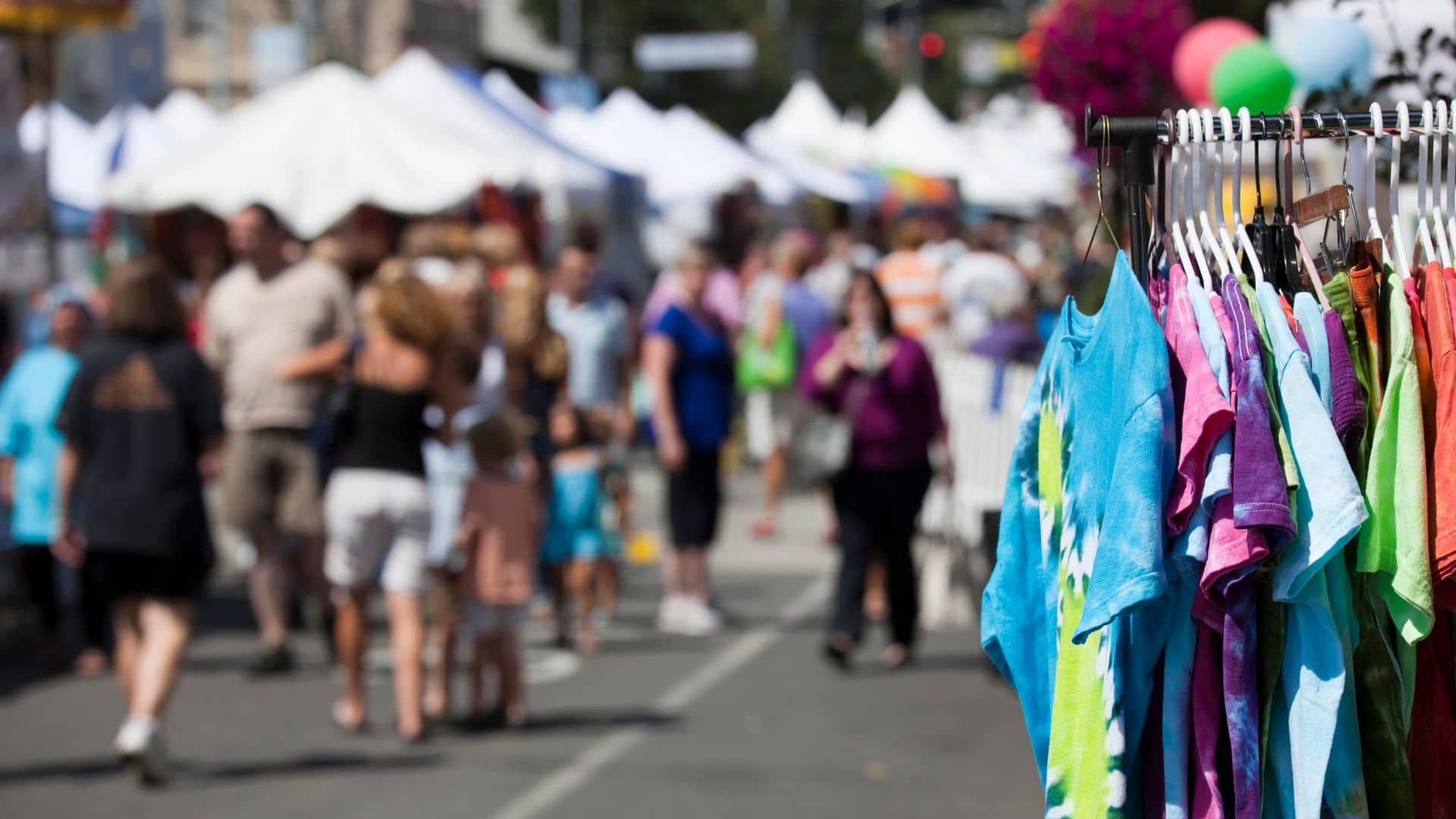 A vibrant craft fair unfolds in the street market scene, with people browsing stalls against a backdrop of colorful tie-dye shirts hanging on a rack. White tents line the bustling street under a clear sky, capturing the essence of a perfect weekend outing.