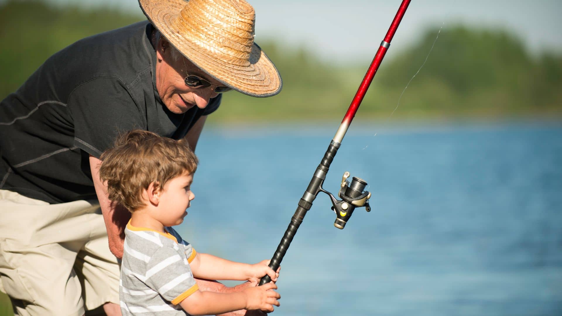 An older man in a straw hat and sunglasses assists a young child with a fishing rod at the lake, resembling a peaceful scene from a campground. The child, donning a striped shirt, is intently focused on their task against the backdrop of shimmering water and fuzzy greenery.