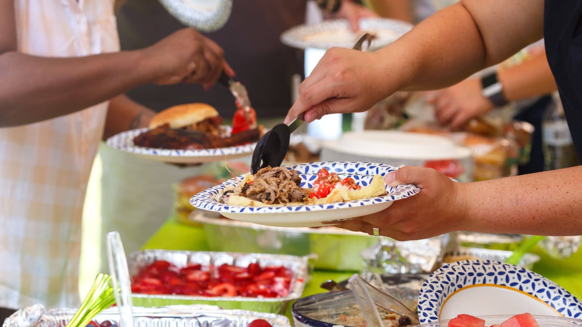 Guests eagerly serve themselves at the potluck dinner, hands balancing plates brimming with pulled pork, salads, and baked beans. A campground vibe lingers as a strawberry dessert awaits in the background amidst friendly chatter and delicious aromas.