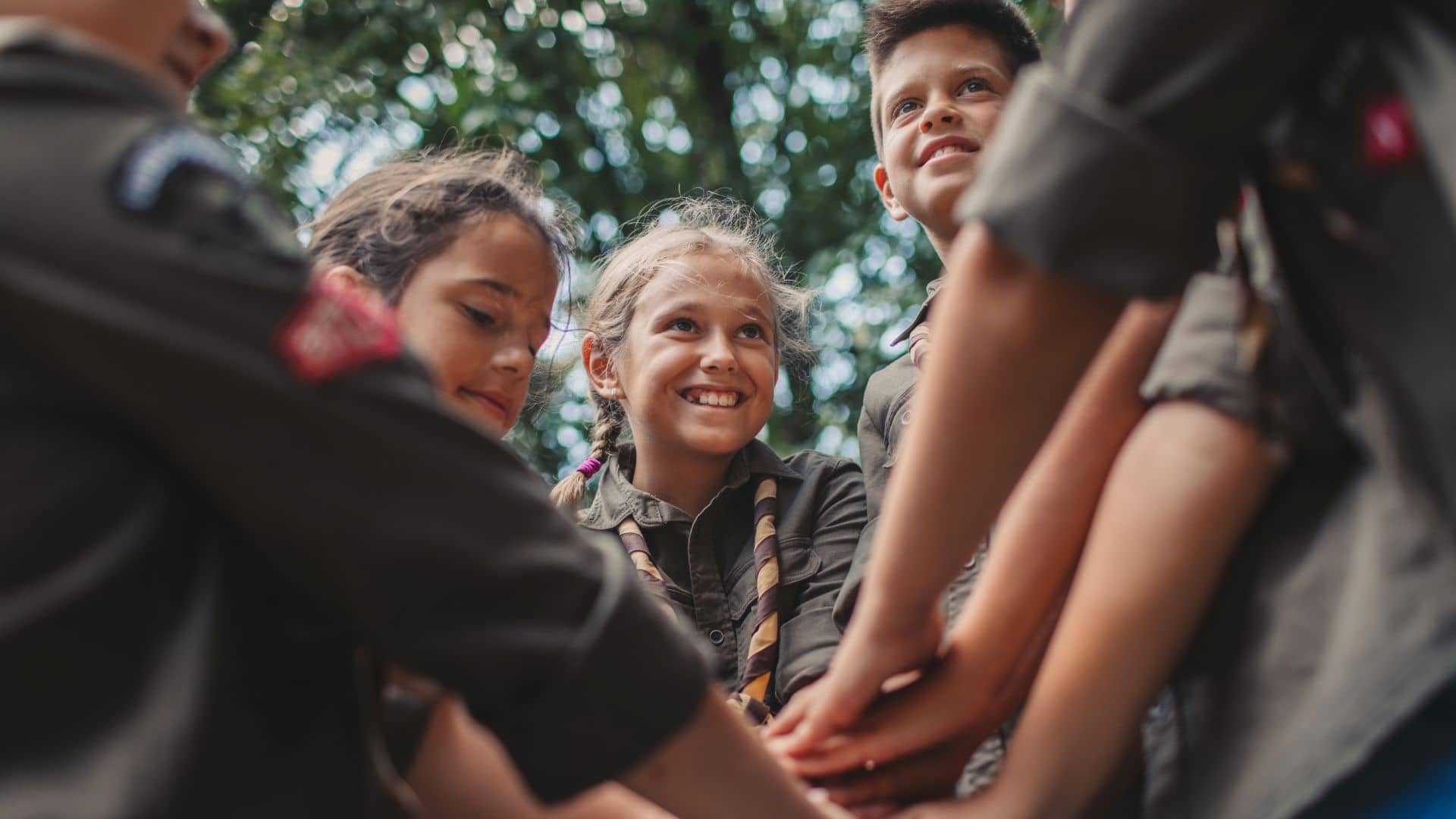 A group of children in scout uniforms huddles together, smiling and stacking their hands in the middle as a sign of teamwork and unity. They stand outdoors at Spacious Skies Campgrounds, with trees visible in the background, embodying the spirit of Scouting America Groups.