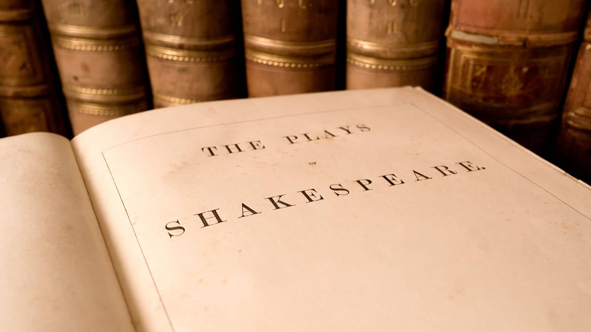 An old book titled "The Plays of Shakespeare" is open, with the title prominently displayed, reminiscent of an Upstate Shakespeare Festival. The background features a row of worn, thick books with leather spines on a shelf.