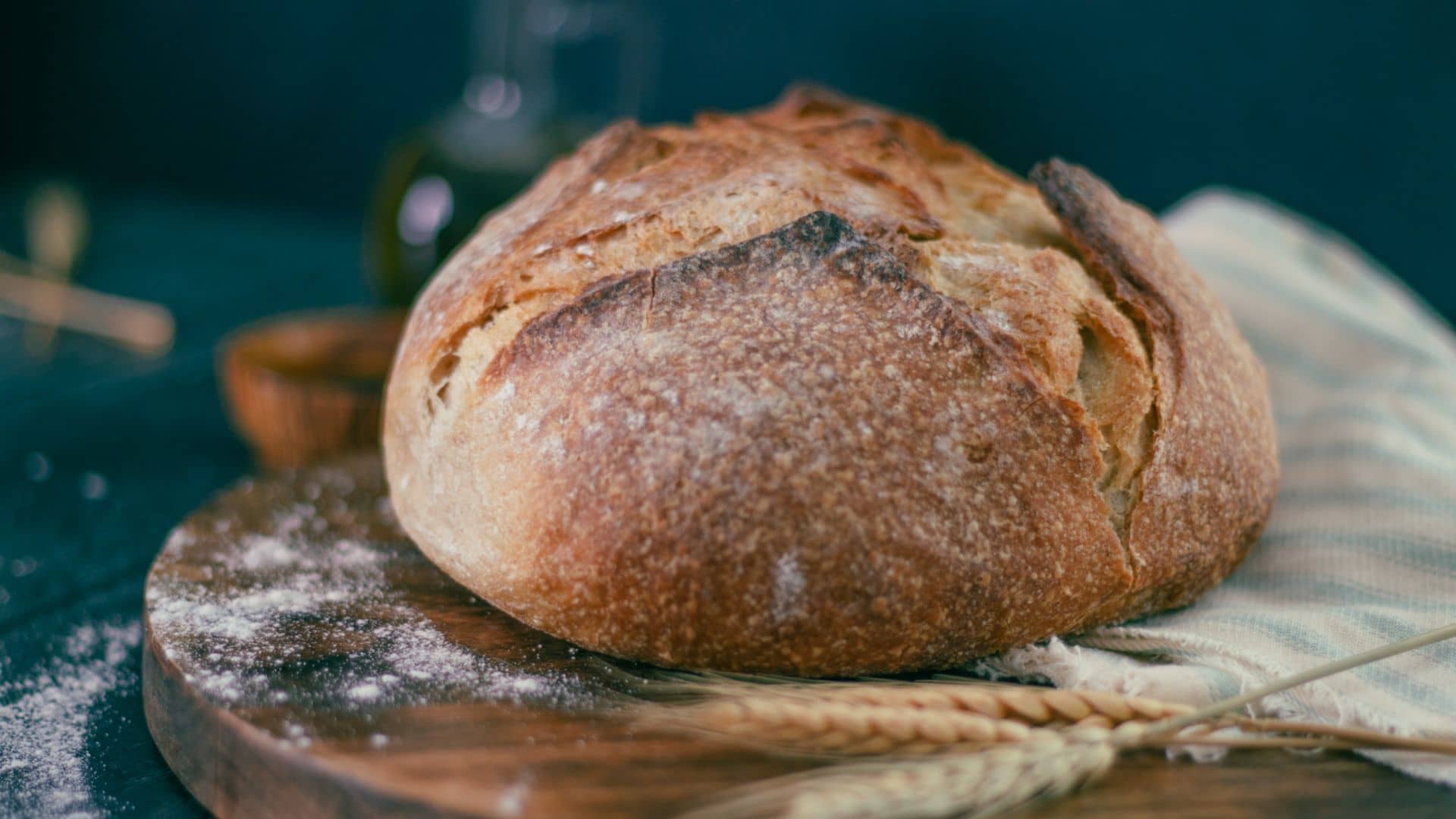 A round loaf of crusty bread sits on a wooden board with a dusting of flour, reminiscent of artisanal offerings at Fretwell Market. Two stalks of wheat rest nearby, with a striped cloth partially covering the board. The background is dark and slightly blurred.