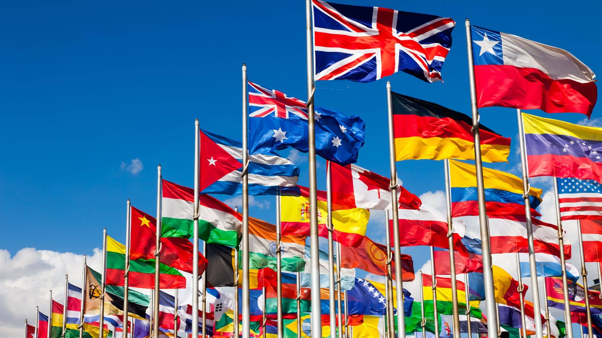 A colorful array of international flags is flying on flagpoles against a bright blue sky with scattered clouds. The vibrant display, reminiscent of an International Festival in Spartanburg, represents global unity with flags arranged closely together.