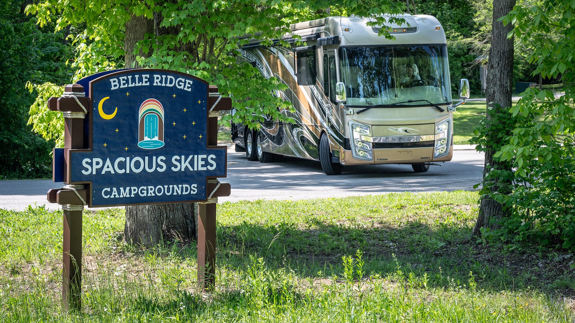 A tan and gold RV parked near a tree-lined road, with a colorful sign in the foreground reading "Belle Ridge Spacious Skies Campgrounds.