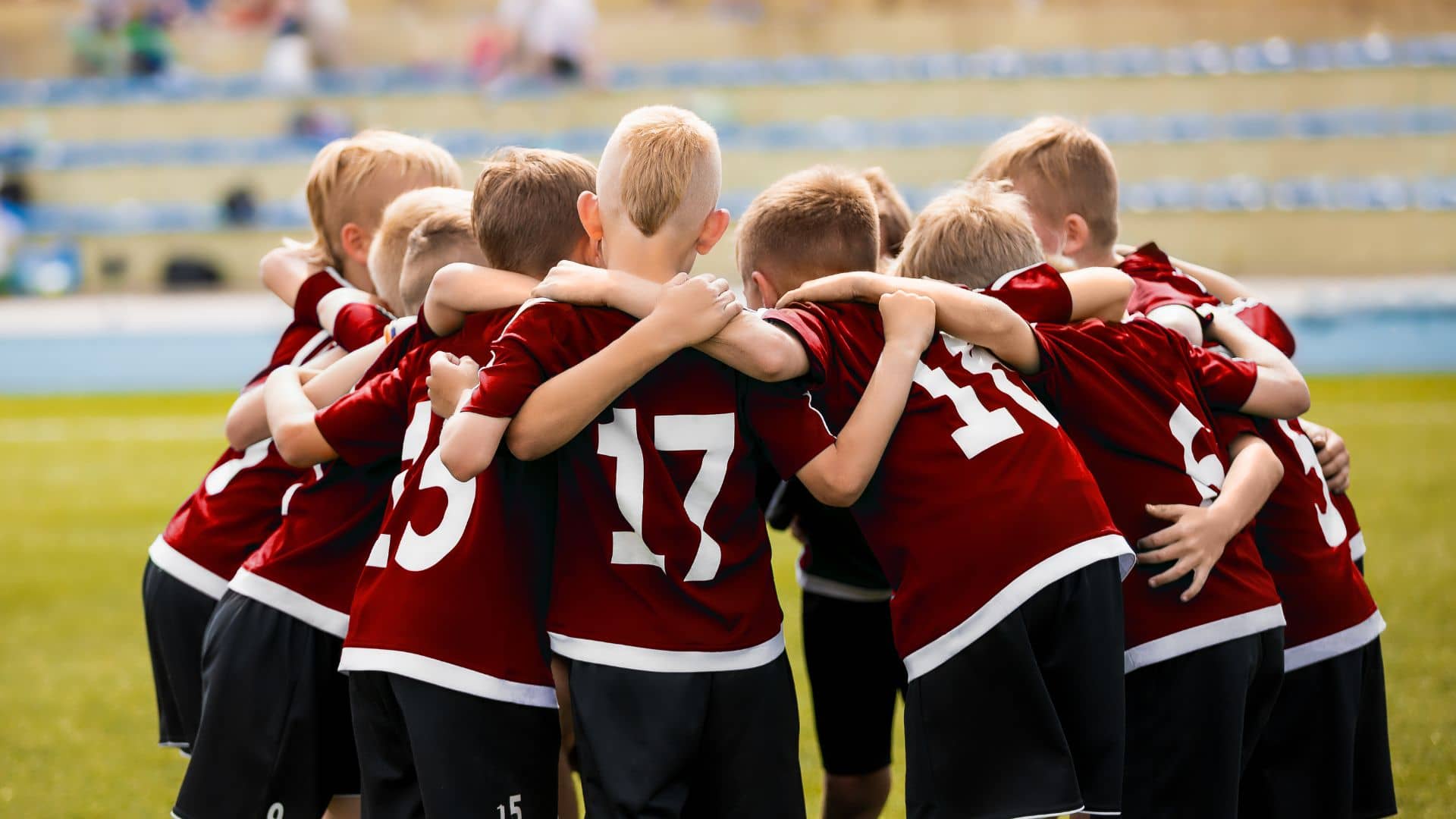 A group of young boys in matching red soccer jerseys huddle together on a grassy field, arms around each other, preparing for a sports tournament. The stands in the background are blurred.