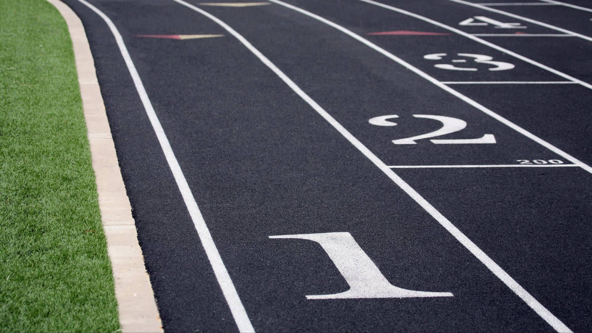 Close-up of a black running track with white lane markings and numbers 1–4, ideal for USATF Junior Olympic events. A strip of green grass borders the left side, capturing the spirit of Track & Field Championship competitions.