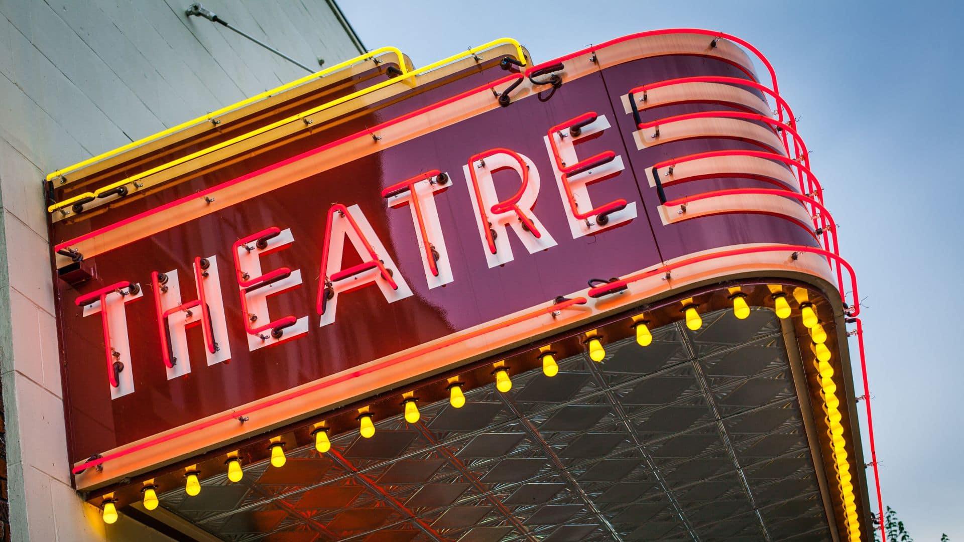 A vibrant red and yellow neon theatre marquee with "THEATRE" lit up, set against a blue sky and glowing bulbs lining the sign, announces the New Play Festival in South Carolina.