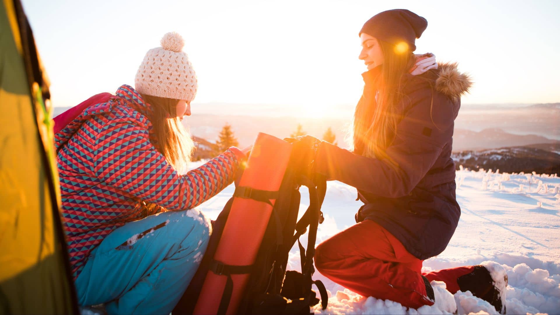 Two people in winter jackets and hats kneel in the snow at sunrise, packing or unpacking gear from a red bag near a tent, enjoying winter camping beneath the spacious skies of Shenandoah, with mountains and trees in the background.