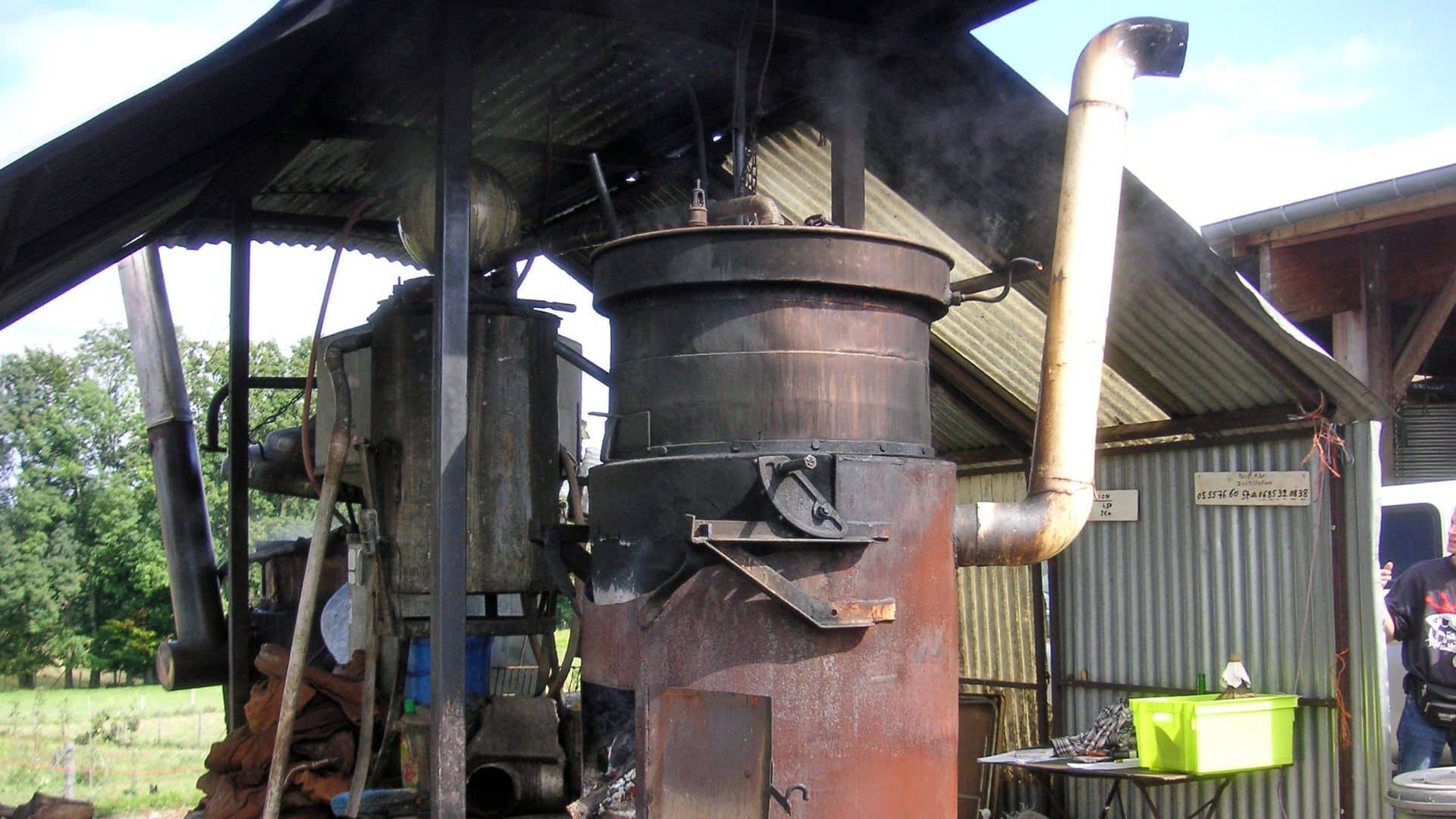 A metal distillation apparatus emits steam under a corrugated metal roof outdoors, with pipes and equipment visible around it.