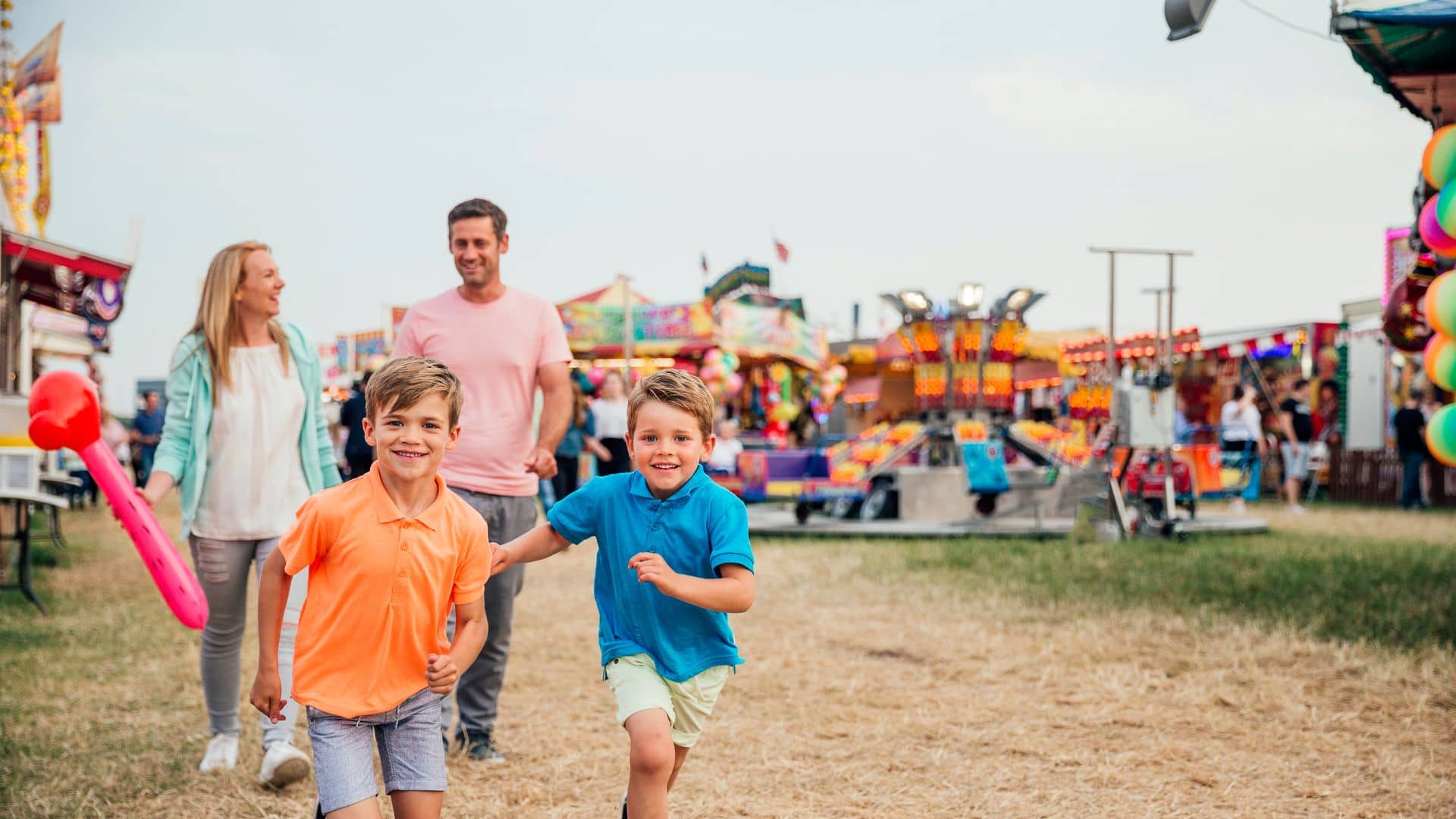 Two children run towards the camera at a colorful outdoor fair, with two adults walking behind them amid various carnival rides and booths.
