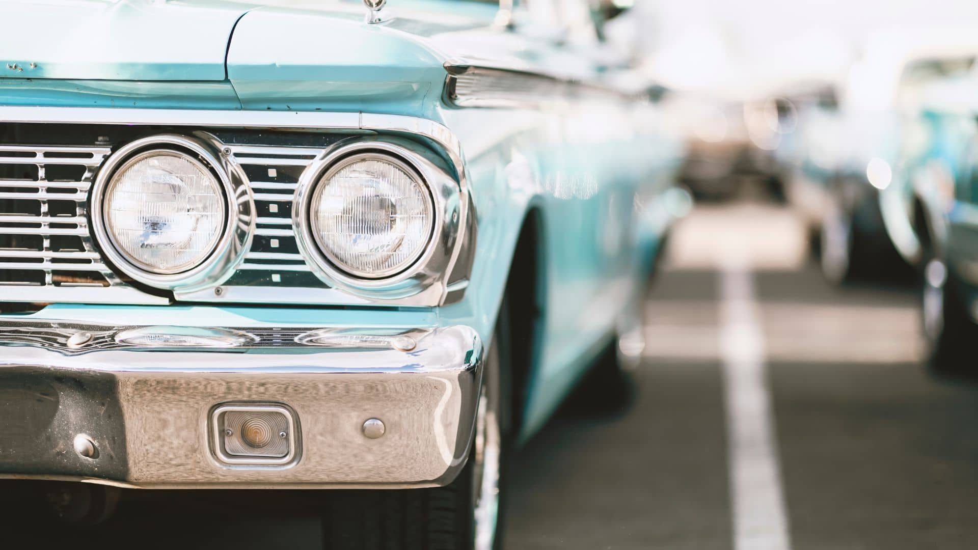 Close-up view of the front left side of a vintage blue car parked in a parking lot, showing headlights, grille, and part of the bumper.