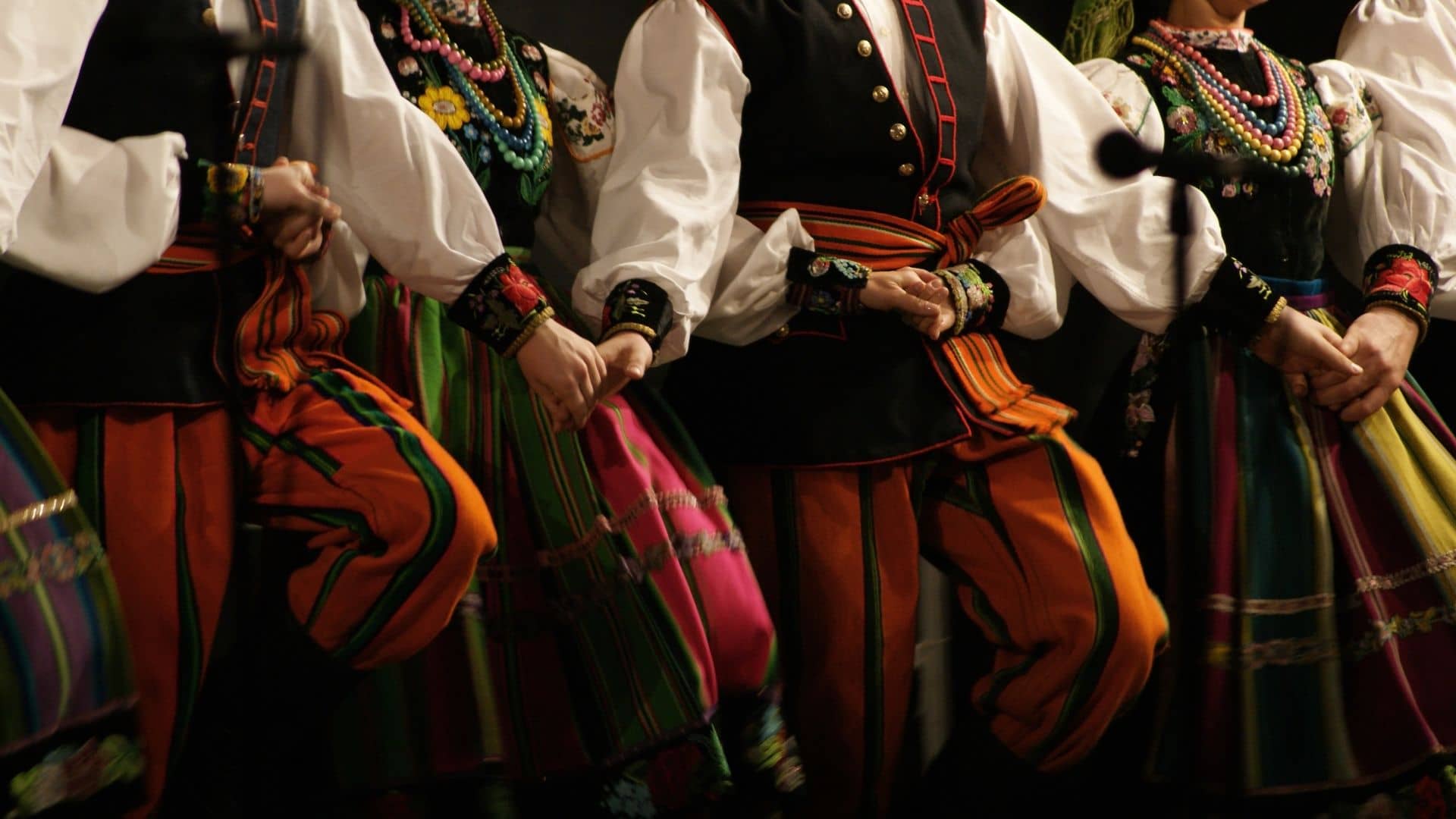 A group of people in traditional folk costumes stand in a line holding hands, wearing colorful striped skirts and pants with decorative vests.