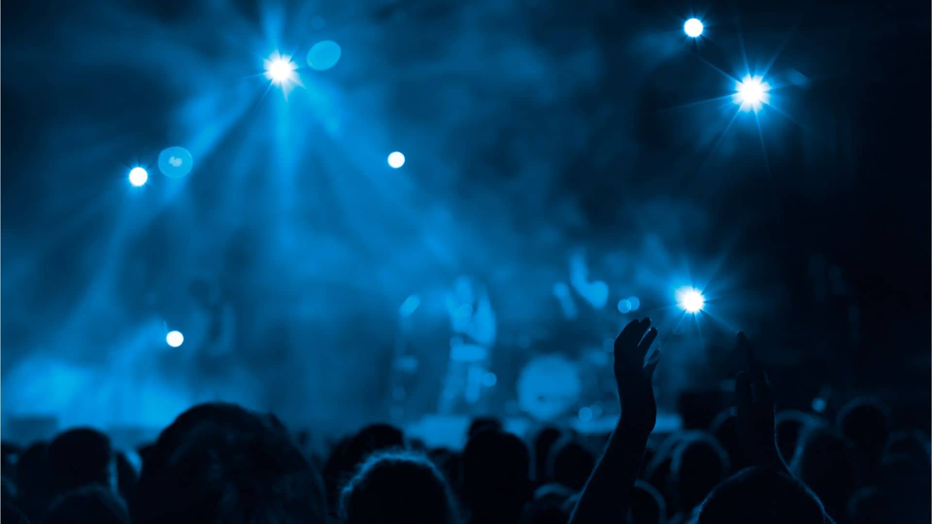 A crowd watches a band perform on stage under blue lighting, with several bright spotlights shining above and raised hands visible in the audience.