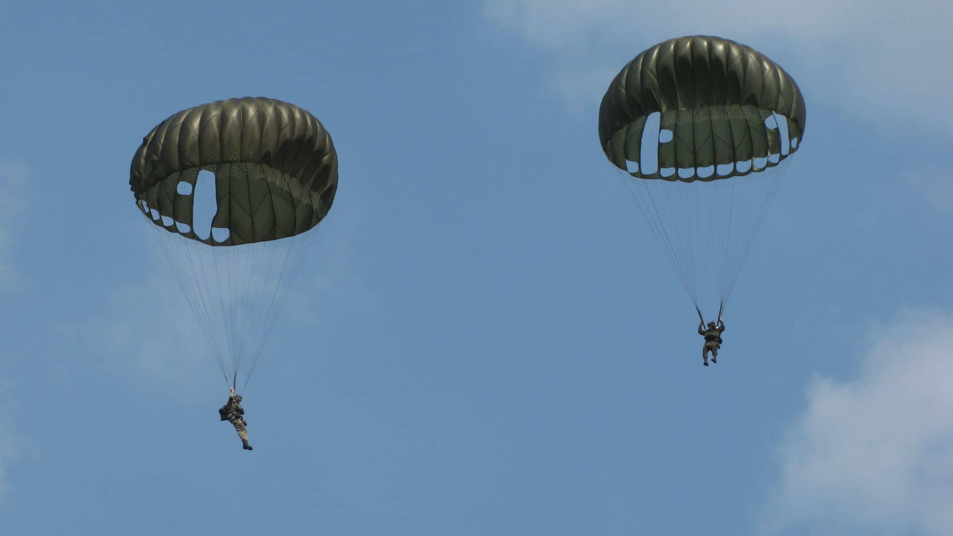 Two people wearing parachutes descend through the sky against a backdrop of blue sky and clouds.