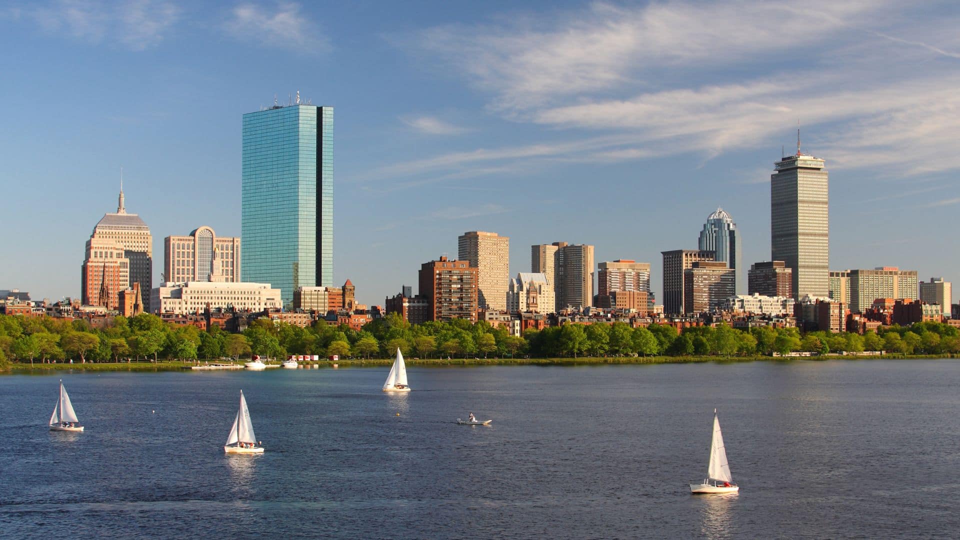 Sailboats on a river with a city skyline and skyscrapers in the background under a blue sky with scattered clouds.