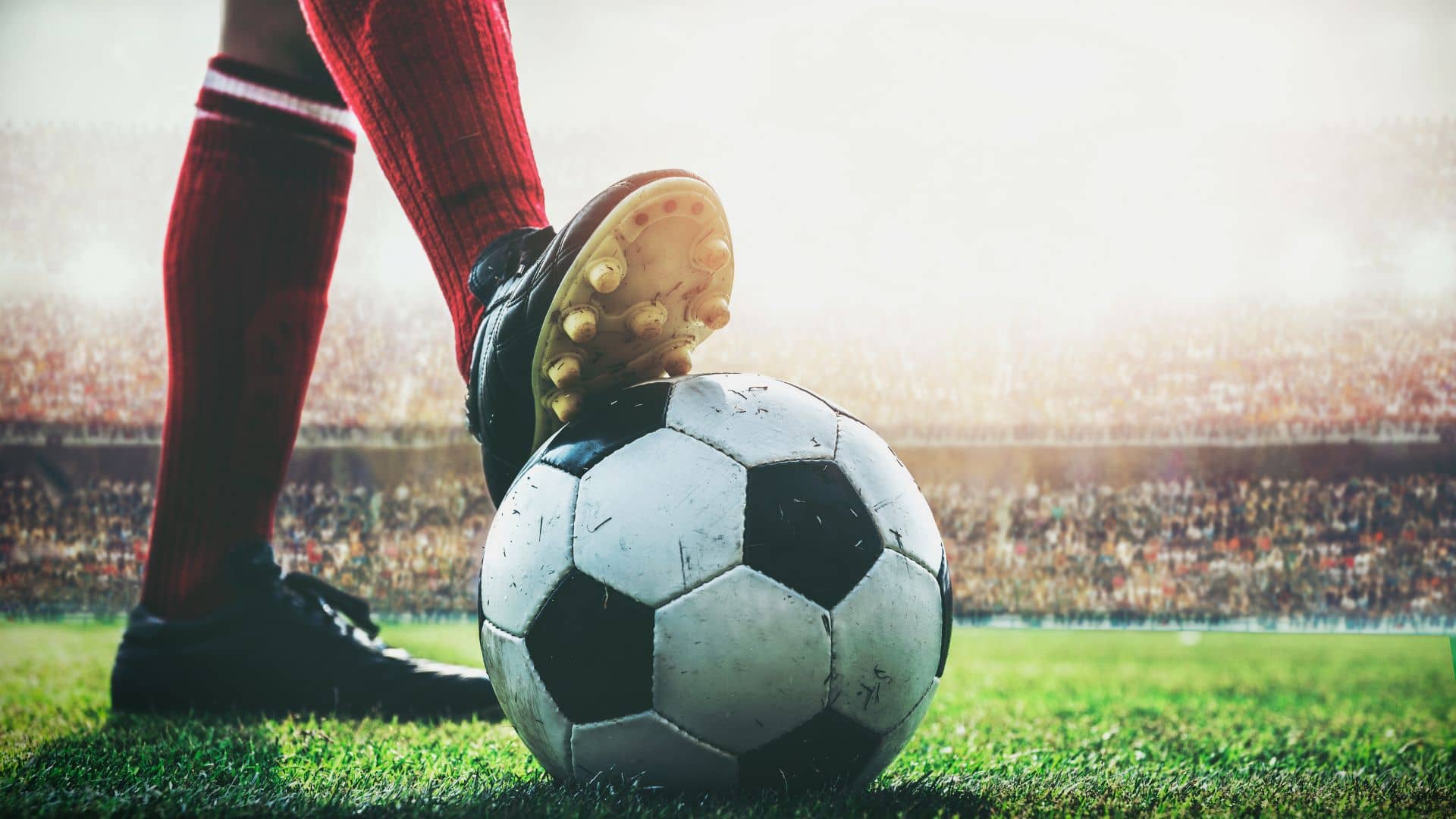 A close-up of a soccer player's Adidas-clad foot resting on a black-and-white ball on a grassy field, with a crowded stadium and bright sunlight in the background during the Savannah Cup.