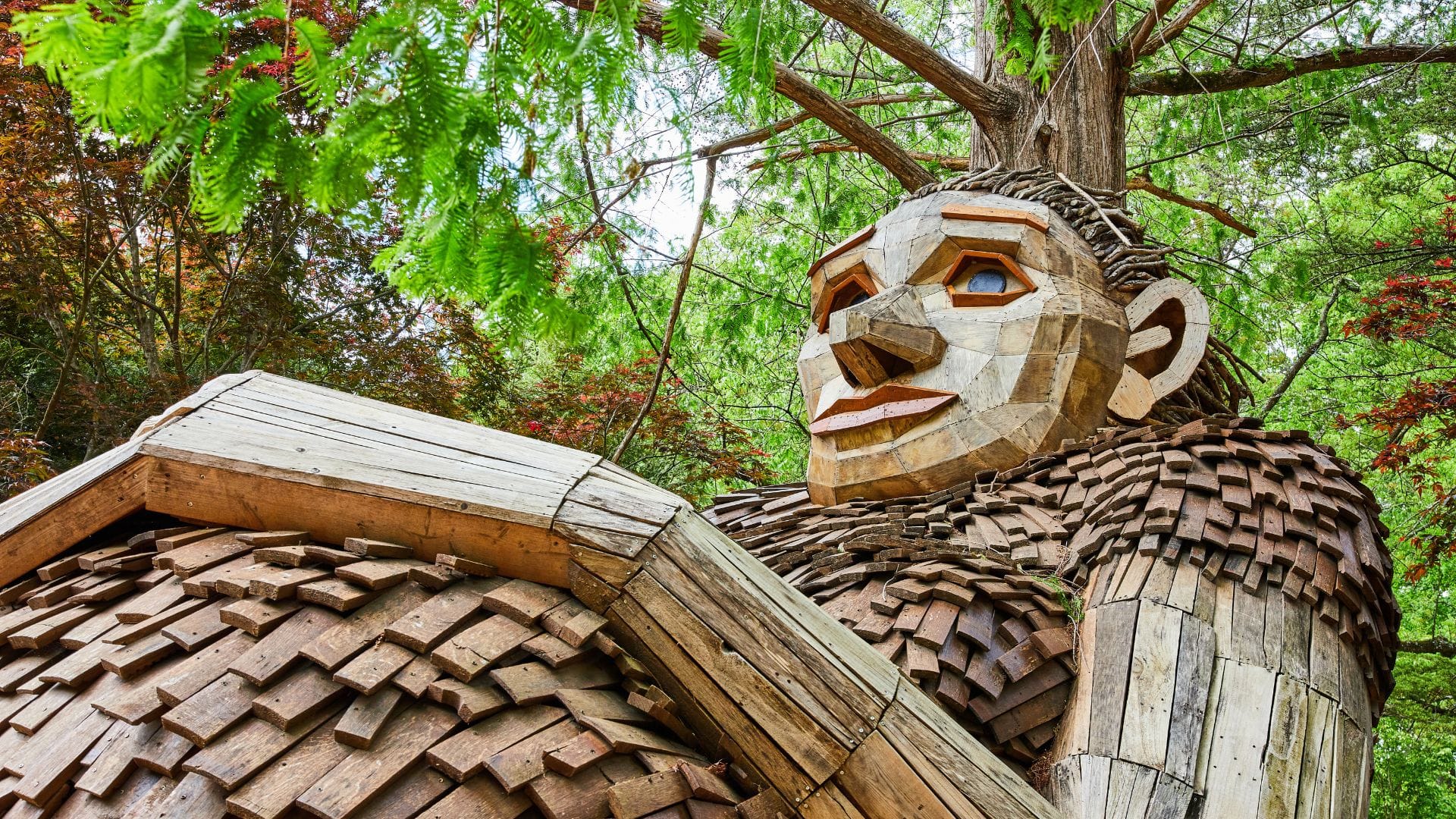 Large wooden sculpture of a giant TROLL with a textured body and shingles stands among green trees at the Wild Center, gazing upward with an expressive face, inviting visitors to join the Save the Humans movement.