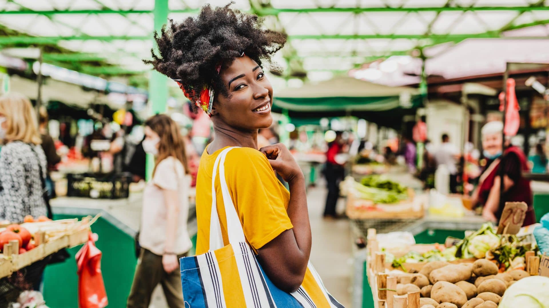 A smiling woman with a striped tote bag stands at the lively Savannah Sunset Market, surrounded by fresh produce and people. She wears a yellow shirt and colorful headband, with vibrant stalls and greenery in the background.
