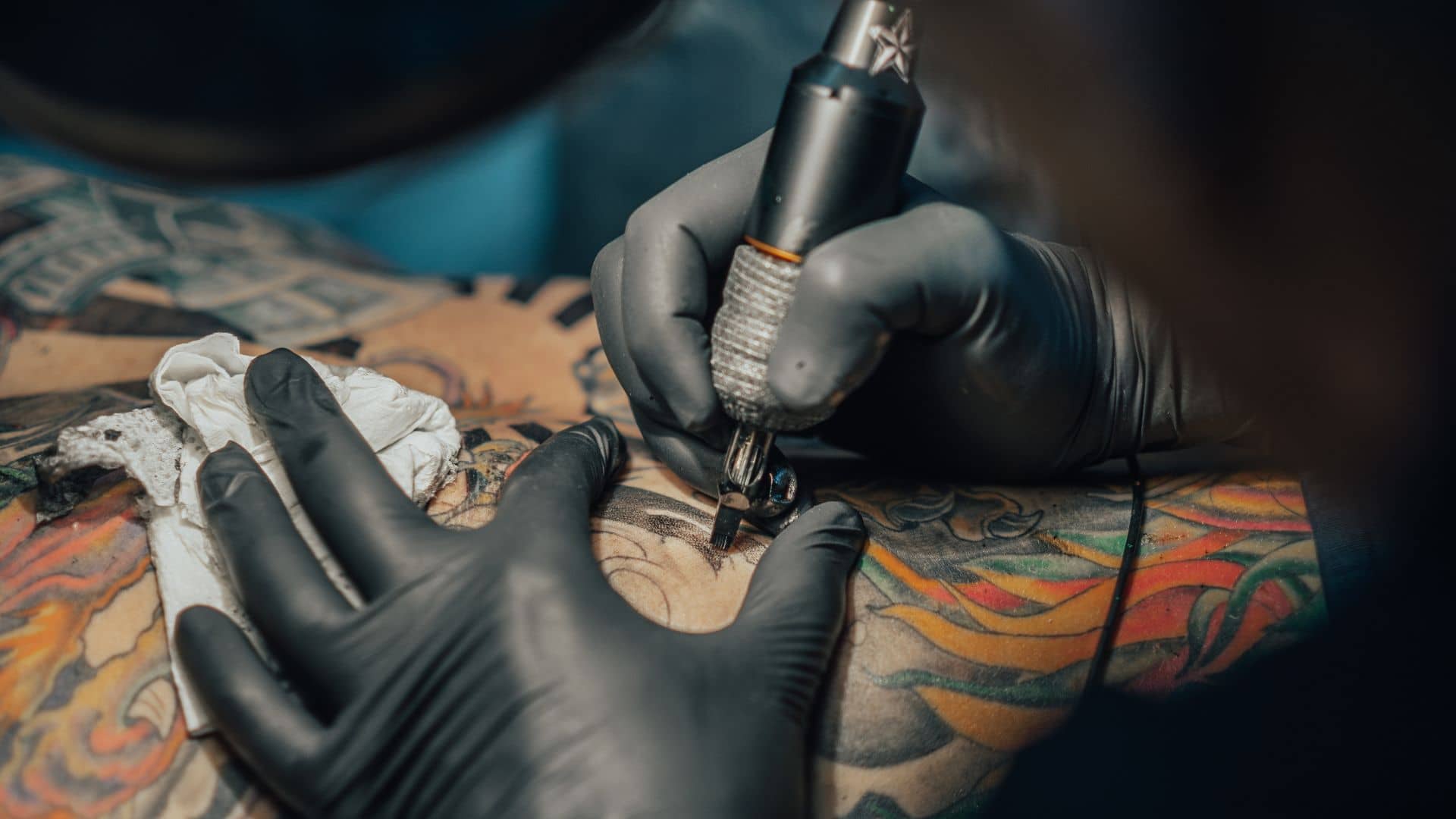 A tattoo artist at the Savannah Tattoo Arts Festival, wearing black gloves, uses a tattoo machine to ink a colorful design on a person's skin while stretching it, with paper towels nearby.